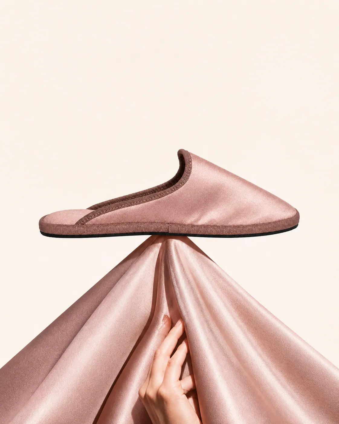 A blush satin Le Furlé slipper balanced on gathered satin, a hand holding the fabric from below