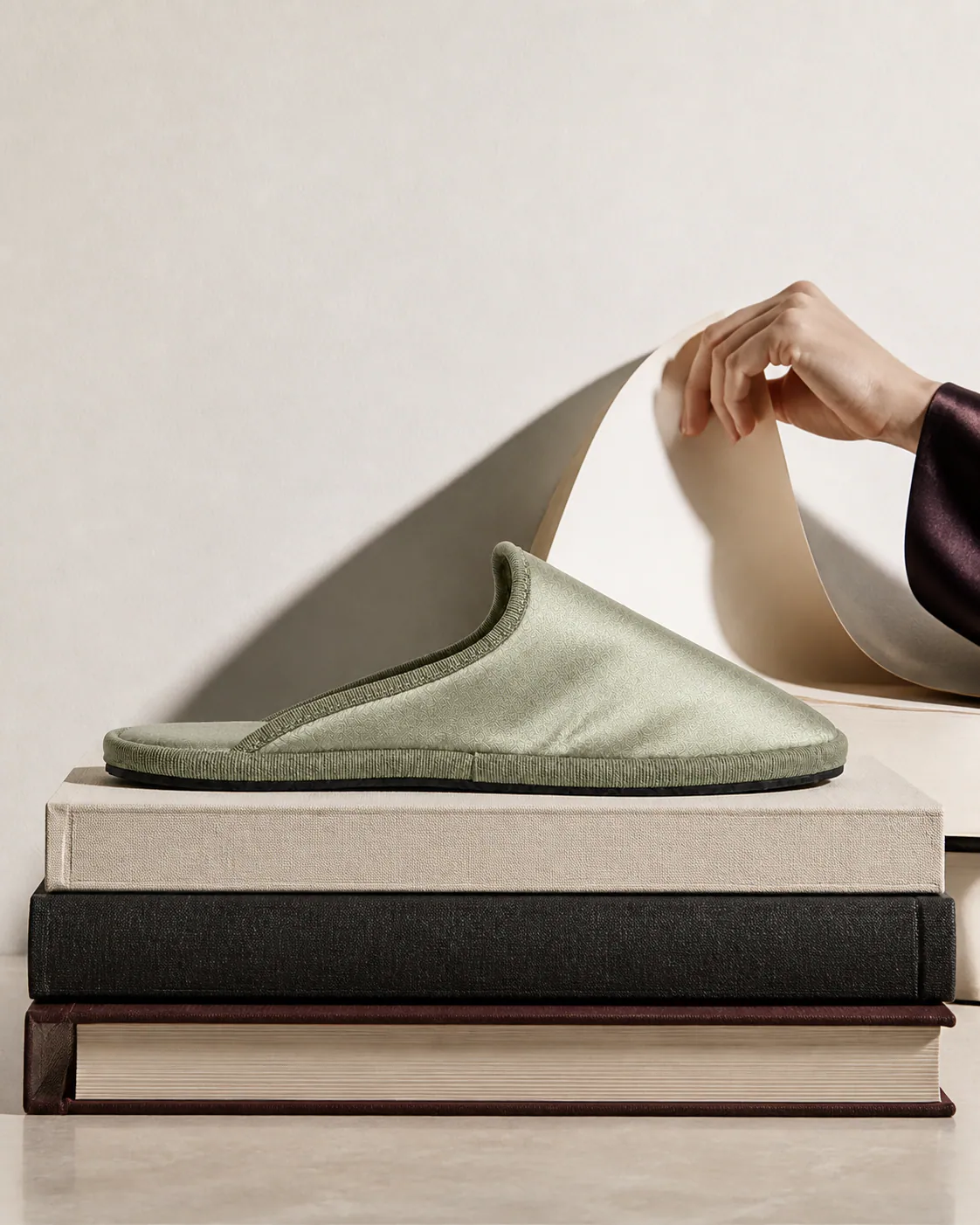 A sage Le Furlé slipper resting on a stack of cloth-bound books while a hand lifts a curl of paper behind it